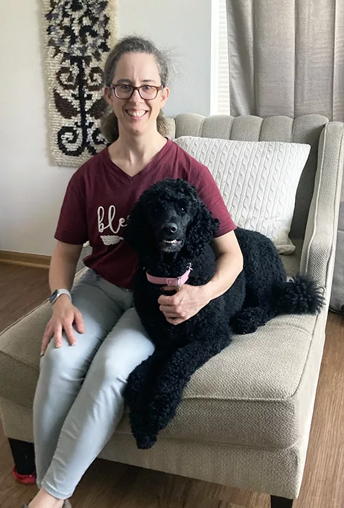 Woman and black standard poodle sitting on sofa together and smiling