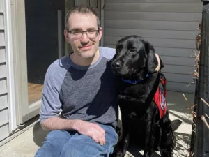 man sitting on front step with arm around black Lab dog wearing red service cape