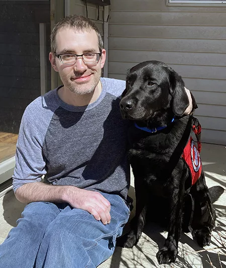 man sitting on front step with arm around black Lab dog wearing red service cape