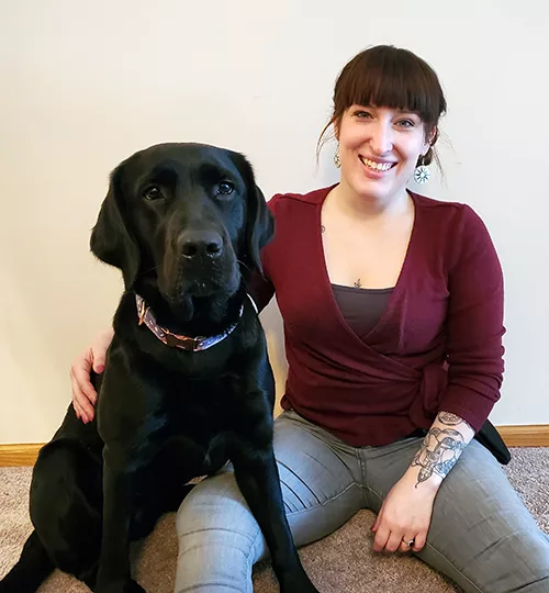 woman sitting on carpeted floor with arm around black Lab dog while smiling at camera