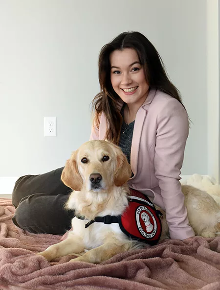 young woman in pink blazer sitting with Golden Retriever service dog wearing red Can Do Canines cape together on mauve blanket on floor