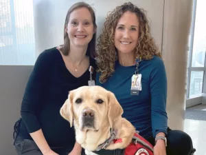 Two women kneeling down to put arms around yellow Lab service dog wearing red Can Do Canines vest
