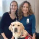 Two women kneeling down to put arms around yellow Lab service dog wearing red Can Do Canines vest