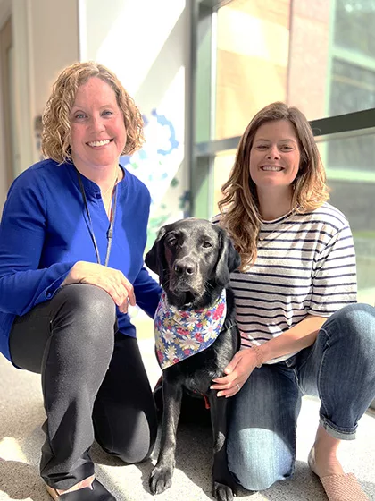 Two women kneeling in business hallway with black Lab dog between them, all smiling at the camera