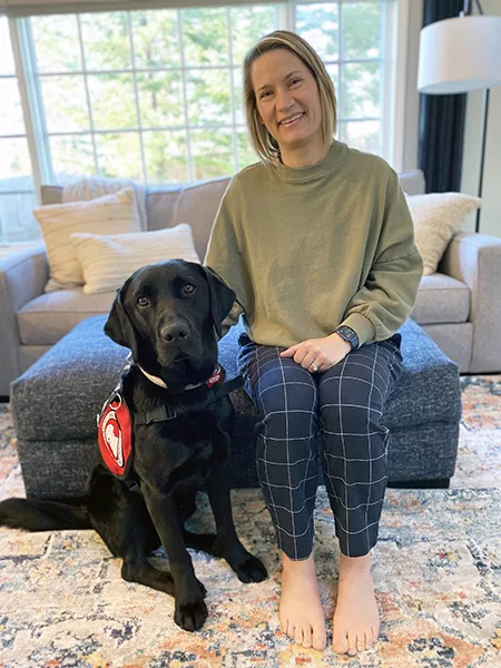 woman sitting on ottoman in living room with black Lab dog wearing service cape sitting next to her on floor, with both looking at the camera