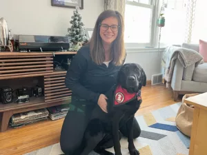 woman kneeling in living room with arm around black Lab that is wearing a red service cape