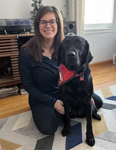 woman kneeling in living room with arm around black Lab that is wearing a red service cape