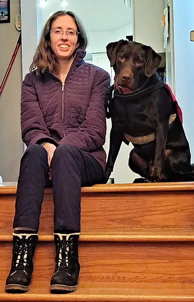 woman and chocolate Lab dog sit together at top of inside stairs smiling at camera