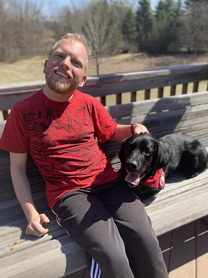 man sitting on deck bench with black Lab service dog sitting beside him