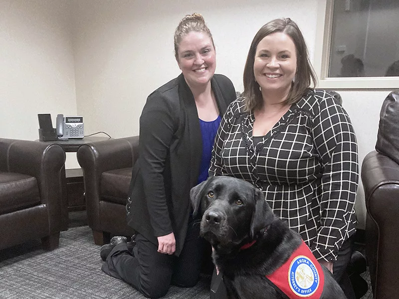 two women kneeling on carpeted floor with black Lab service dog sitting in front of them
