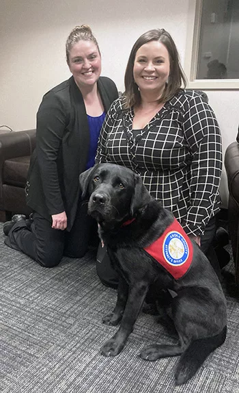 two women kneeling on carpeted floor with black Lab service dog sitting in front of them