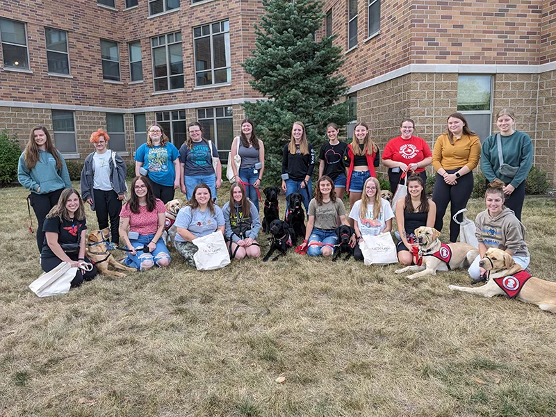 group of about 20 female college students standing outside college building on lawn with about 12 service dogs