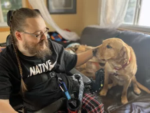 man sitting in wheelchair looking at and putting his hand on a yellow Lab dog sitting on couch near him