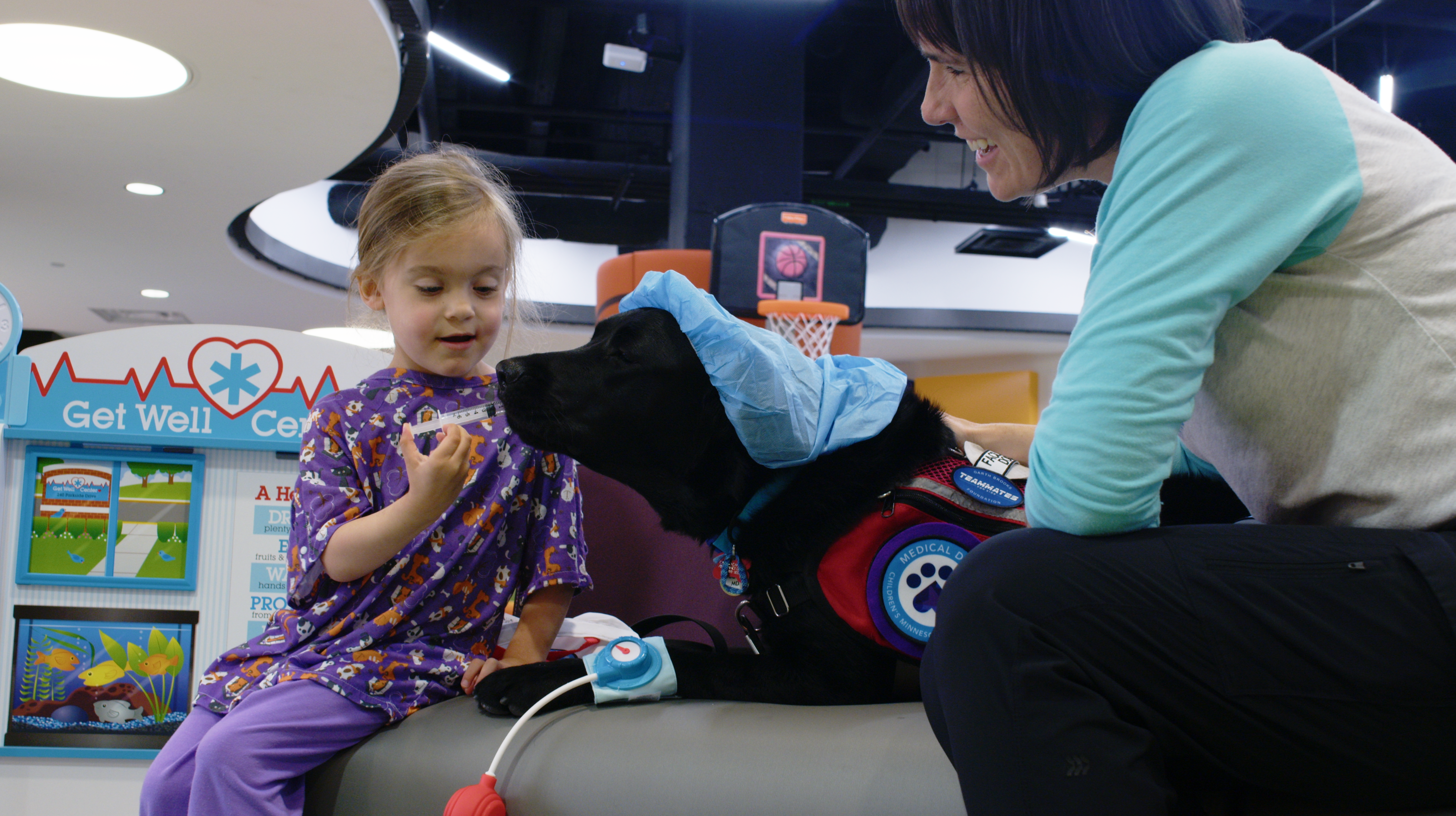 young girl putting a fake syringe into the mouth of a black Lab service dog wearing a hospital scrubs cap