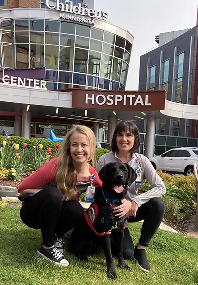 two woman crouching outside on lawn with arms around black Lab service dog between them and all are smiling at camera