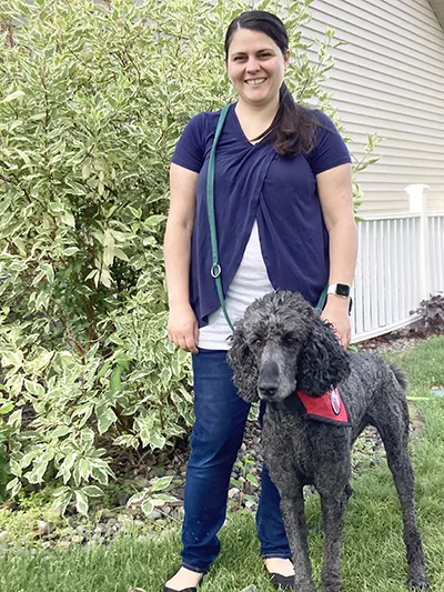 woman standing outside home with gray Standard Poodle wearing red service cape standing beside her