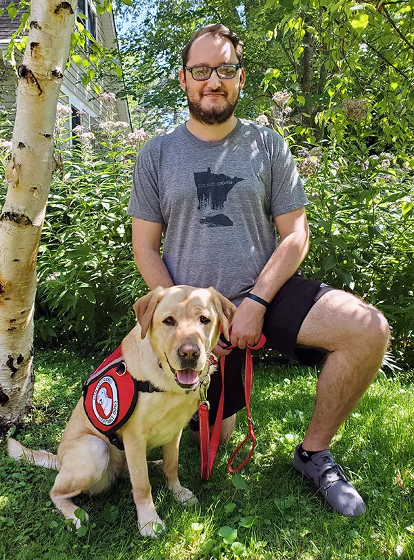 man kneeling on grass with yellow Lab dog wearing Can Do Canines service vest sitting next to him, both are looking at the camera