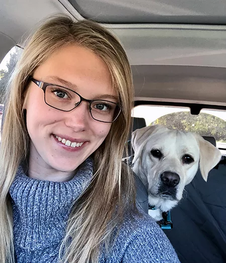 young woman with blond hair and glasses smiling for selfie with yellow Lab in back seat of car behind her