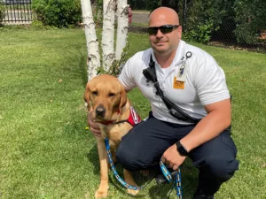 man in EMT uniform crouching on grass with arm around yellow Lab service dog