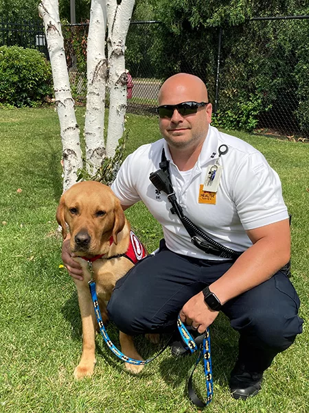 man in EMT uniform crouching on grass with arm around yellow Lab service dog