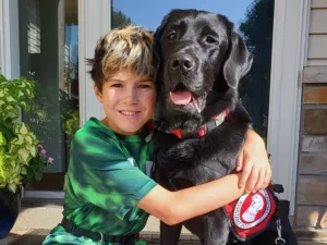 boy sitting on outside steps with arms around black Lab service dog, and both are smiling at the camera