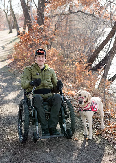 man sitting in wheelchair with yellow Lab dog wearing red service cape with front paws up on man's lap while they are on a path near a river