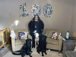 woman standing in living room with two black Labs and one yellow Lab in front of collage of dogs in shape of "100" on wall
