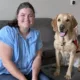 woman sitting in living room near yellow Lab dog with red service cape, and both are smiling at camera