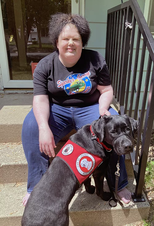 woman sitting on front steps of home with black Lab service dog wearing red Can Do Canines cape standing between her legs