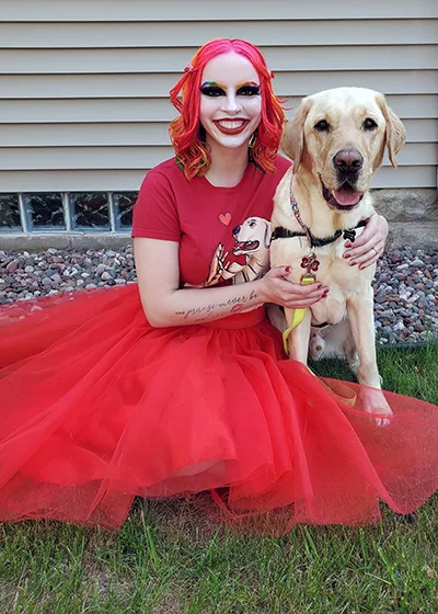 woman wearing red shirt and red tulle skirt sitting on grass with hand on yellow Lab service dog wearing red Can Do Canines cape