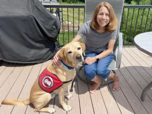 woman sitting on sunny deck with hand on yellow Lab service dog wearing red Can Do Canines cape