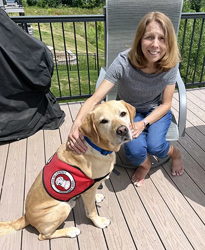 woman sitting on sunny deck with hand on yellow Lab service dog wearing red Can Do Canines cape