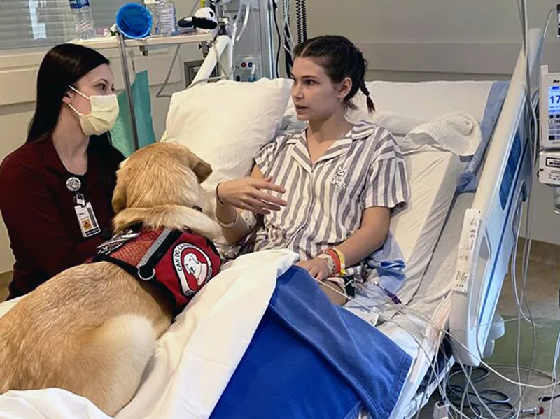 teenage girl sitting in hospital bed, talking to staff member while yellow Lab service dog wearing Can Do Canines red cape lies on bed