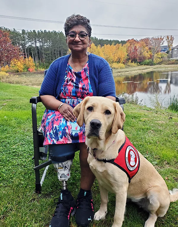 woman sitting in wheelchair outside near pond with yellow Lab sitting in front of her, wearing red Can Do Canines service cape