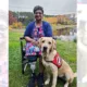 woman sitting in wheelchair outside near pond with yellow Lab sitting in front of her, wearing red Can Do Canines service cape