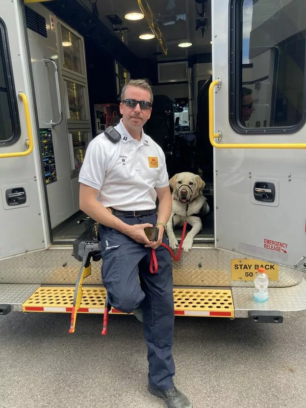 paramedic sitting on opened back of ambulance with yellow Lab service dog lying next to him with both smiling at camera