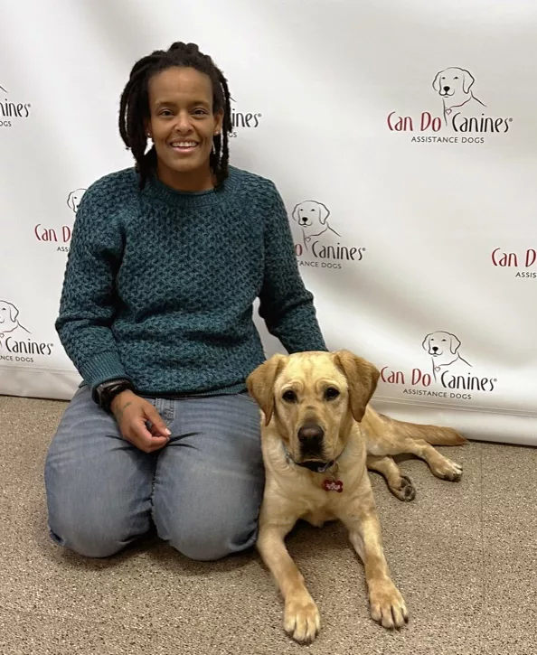 woman kneeling next to yellow Lab that is lying on floor in front of Can Do Canines logo background