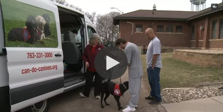 woman handing a leashed black Lab to one of two prison inmates near Can Do Canines van