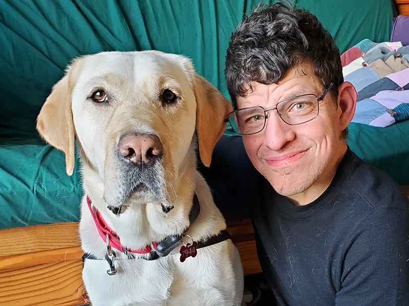 Yellow Lab and man with curly hair and glasses sitting with heads close together smiling at camera