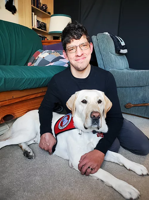 Man sitting on floor with arms around yellow Lab service dog lying next to him; both are looking at the camera