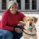 woman crouching down on snow-covered grass outside building with arm around yellow Lab dog
