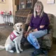 Woman sitting in living room chair with yellow Lab sitting next to her wearing Can Do Canines service vest; both are smiling at camera