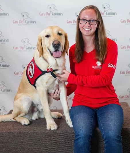 woman wearing red shirt and glasses sitting on platform with yellow Lab service dog sitting next to her in front of backdrop with Can Do Canines logos