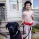 Young girl with curly black hair standing outside front door of home holding leash of black Lab service dog sitting in front of her