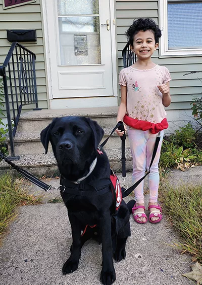 Young girl with curly black hair standing outside front door of home holding leash of black Lab service dog sitting in front of her
