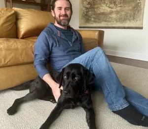 man sitting on a living floor with a black Lab