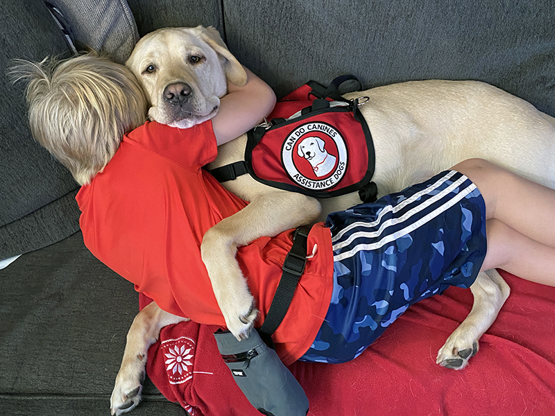 young boy lying on sofa with yellow Lab dog wearing Can Do Canines service cape; both have arms around each other and boy's face is hidden behind dog's face