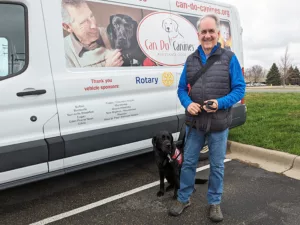 man standing and black Lab wearing red service cape sitting in front of van with siding that says Can Do Canines