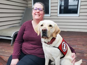 woman sitting on deck outside house with arm around yellow Lab wearing red service cape