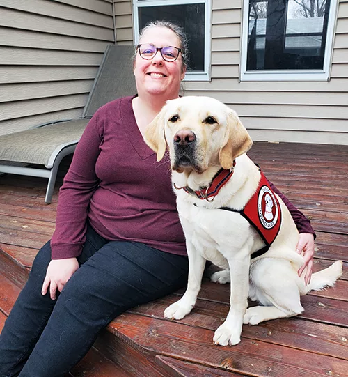 woman sitting on deck outside house with arm around yellow Lab wearing red service cape